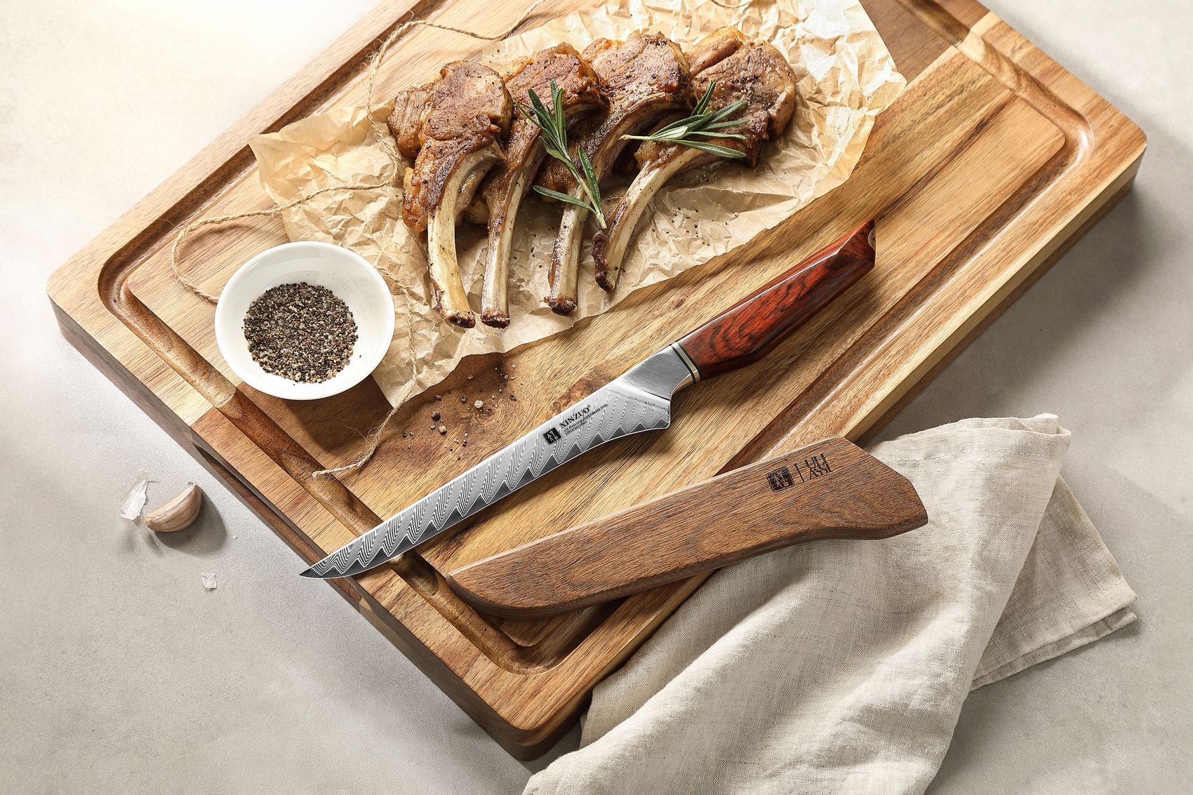 Cooked lamb cutlets with rosemary and cracked pepper on a wood board beside the Damascus boning knife and its wooden saya