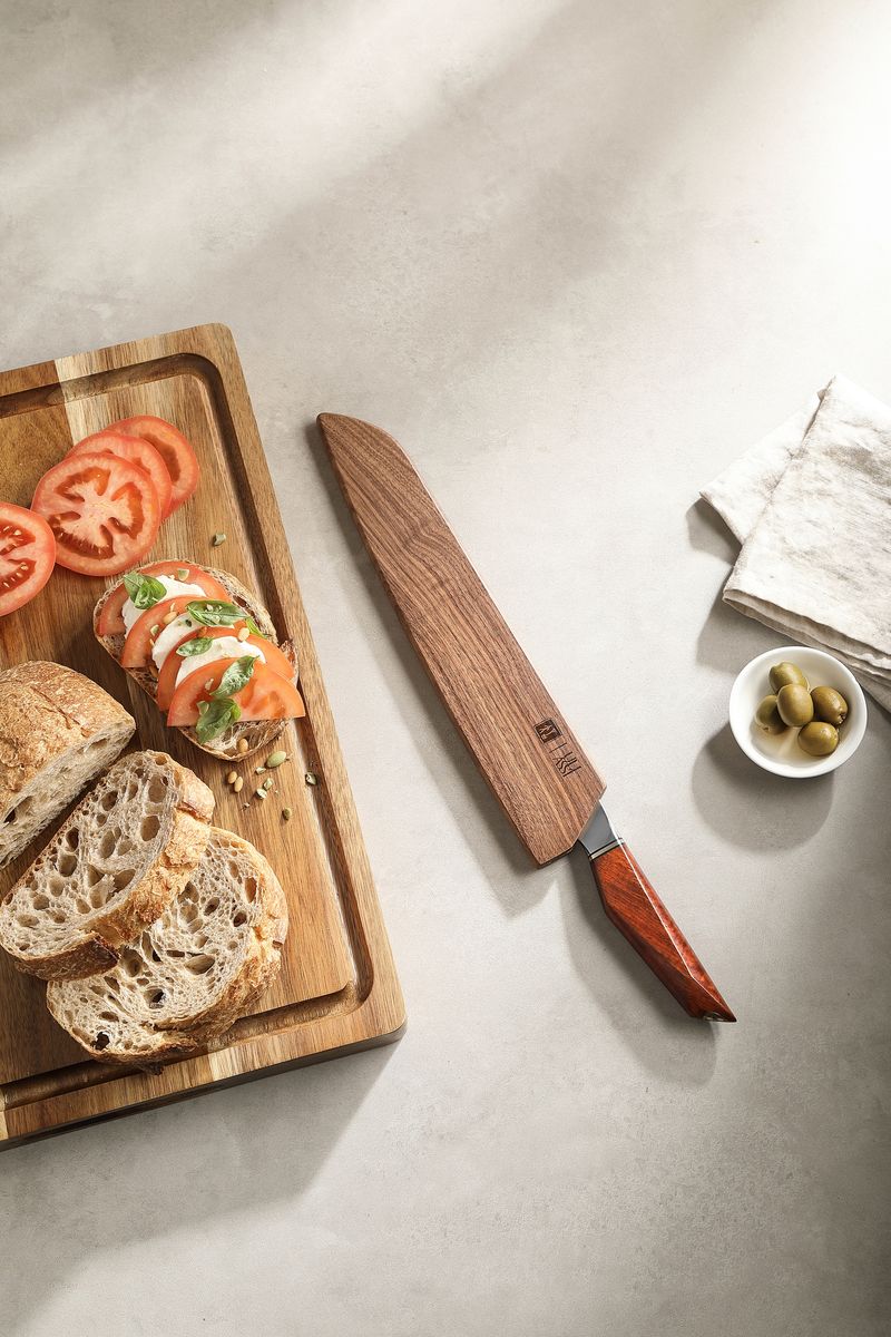 Bread knife sheathed in its wooden saya beside a board of sliced bread and tomato