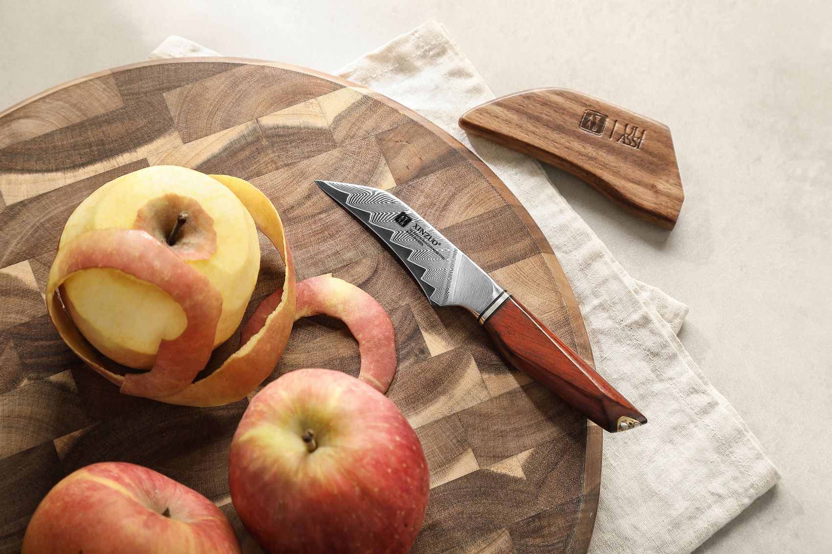 Paring knife on a round end-grain board with peeled apples and a coiled apple peel