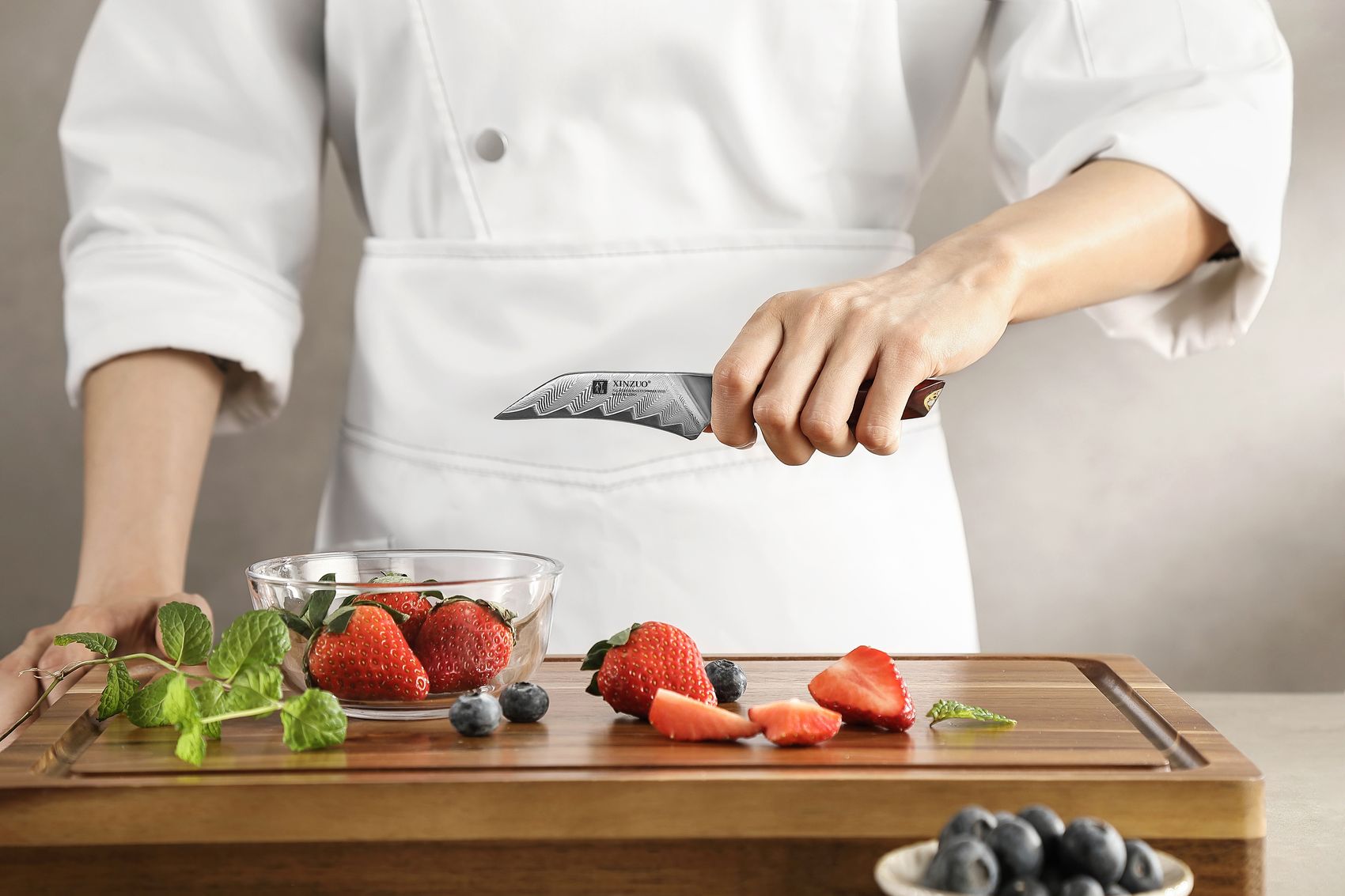 Chef gripping the paring knife while slicing strawberries on a board with blueberries and mint