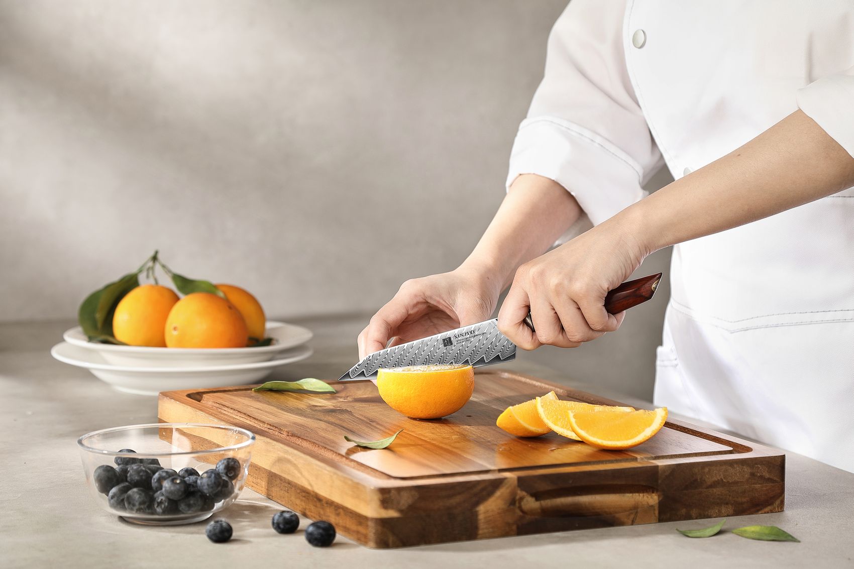 Chef in white jacket slicing an orange on a wooden board with the Damascus utility knife, blueberries and oranges alongside