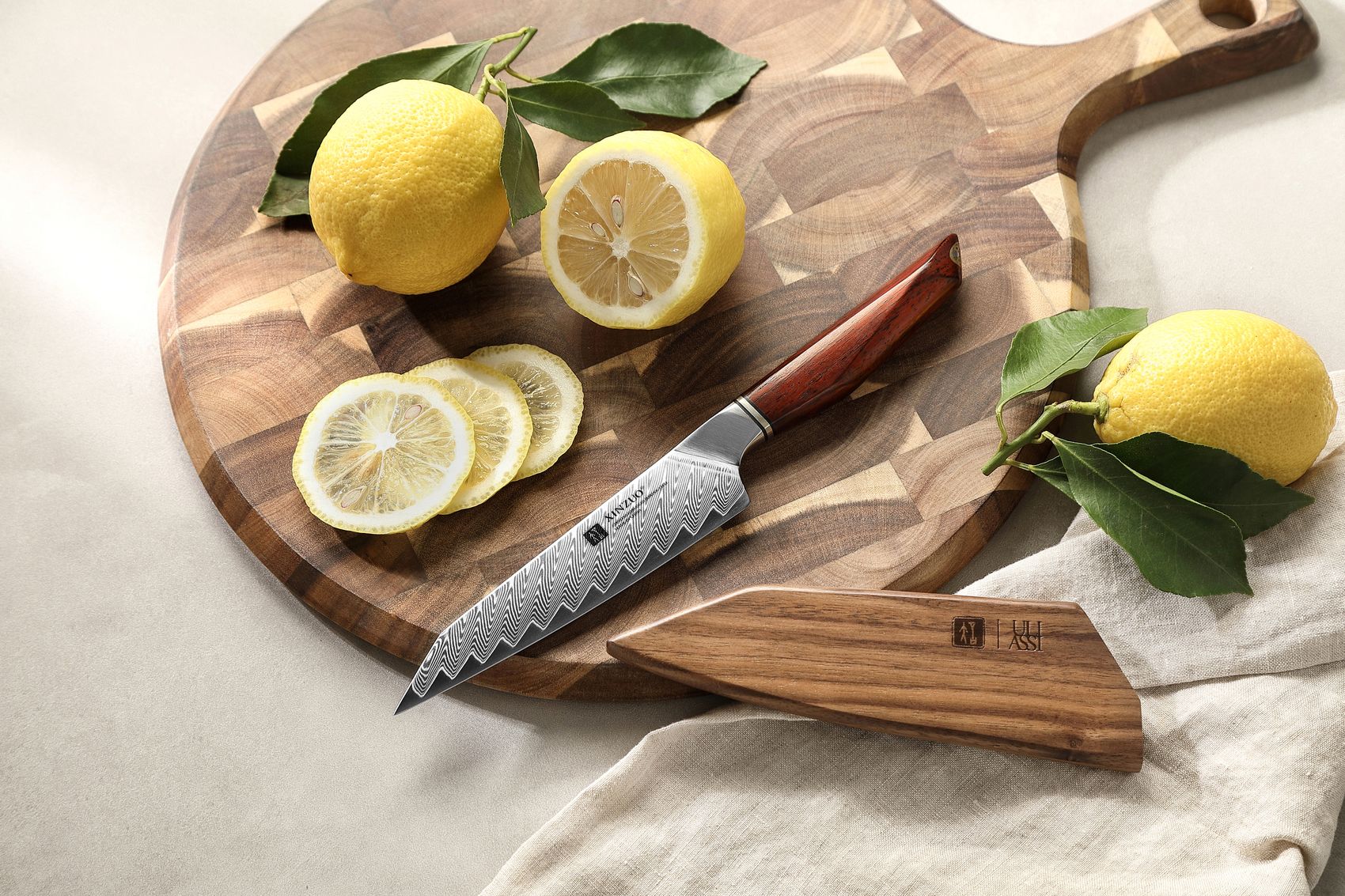 Damascus utility knife on a round wooden board with its matching wooden saya sheath, whole and sliced lemons nearby