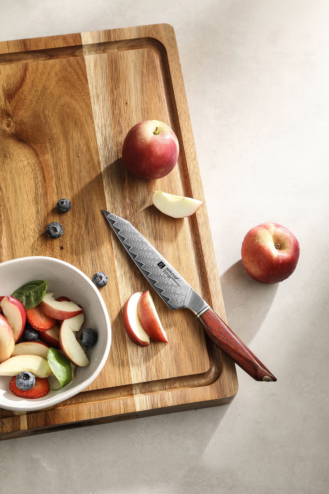 Wooden cutting board with apples, apple slices, blueberries and a fruit-salad bowl, the Damascus utility knife resting alongside
