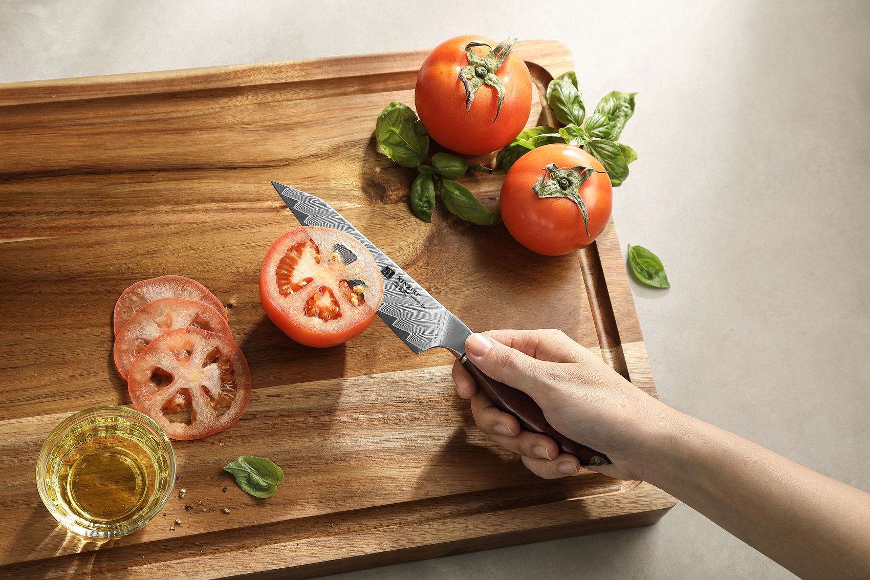 Overhead view of a hand gripping the Damascus utility knife slicing a tomato on a wooden board with basil and olive oil