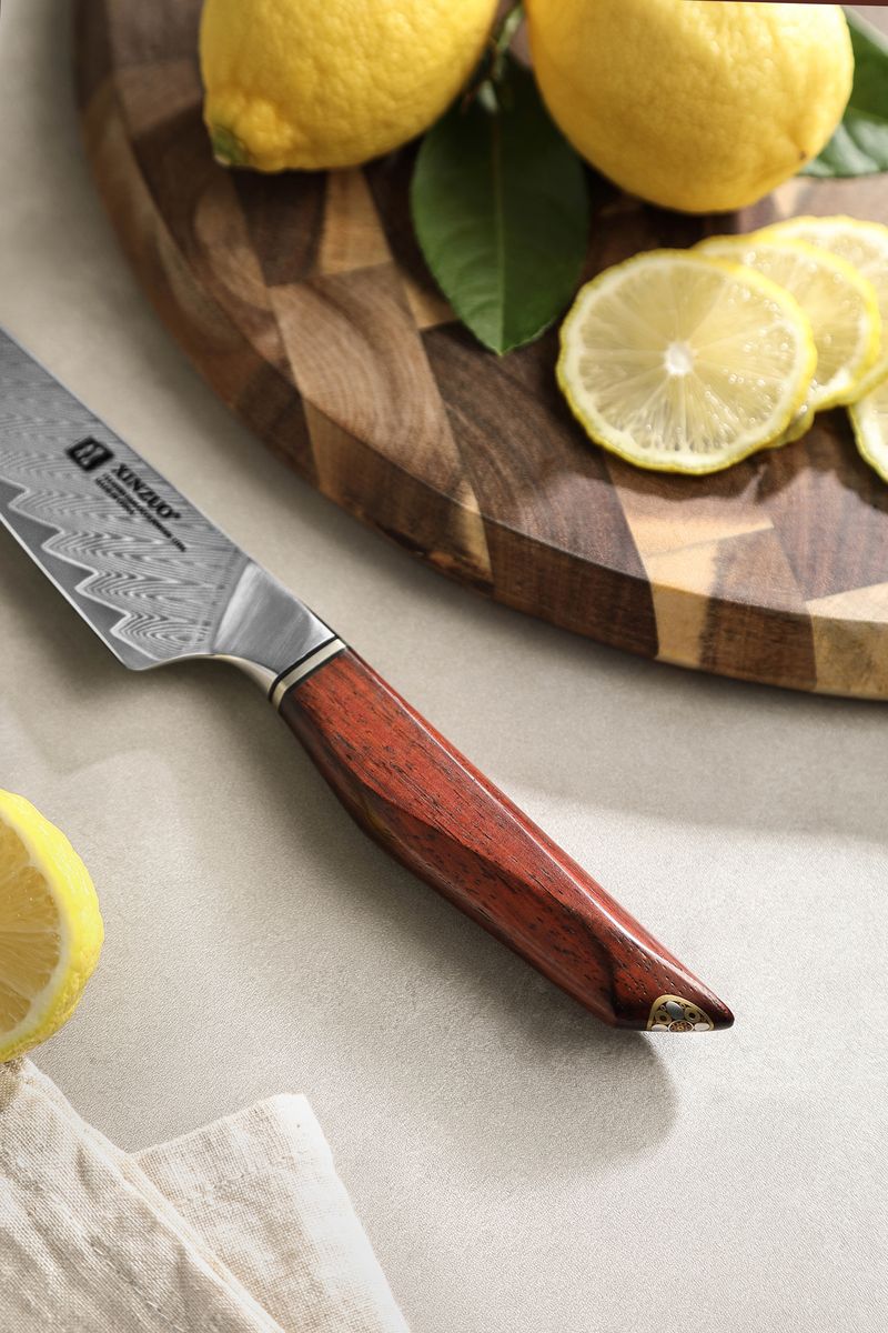 Studio detail of the utility knife showing the Damascus blade pattern, XINZUO etch and rosewood handle beside sliced lemons